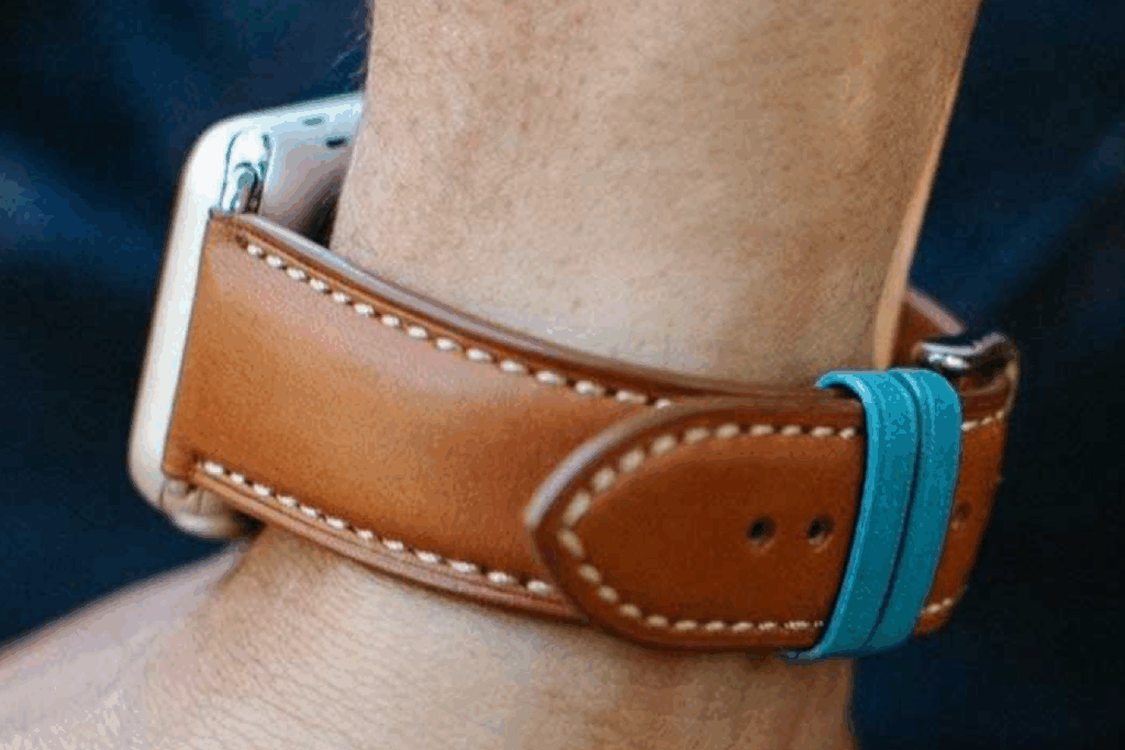 Close-up of a wrist wearing an Apple Watch with a Saddle Tan French premium leather band featuring white stitching and a contrasting blue loop.