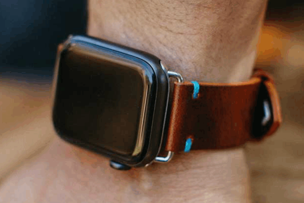 Apple Watch on wrist with a dark brown Italian leather band featuring bright blue stitching accents.
