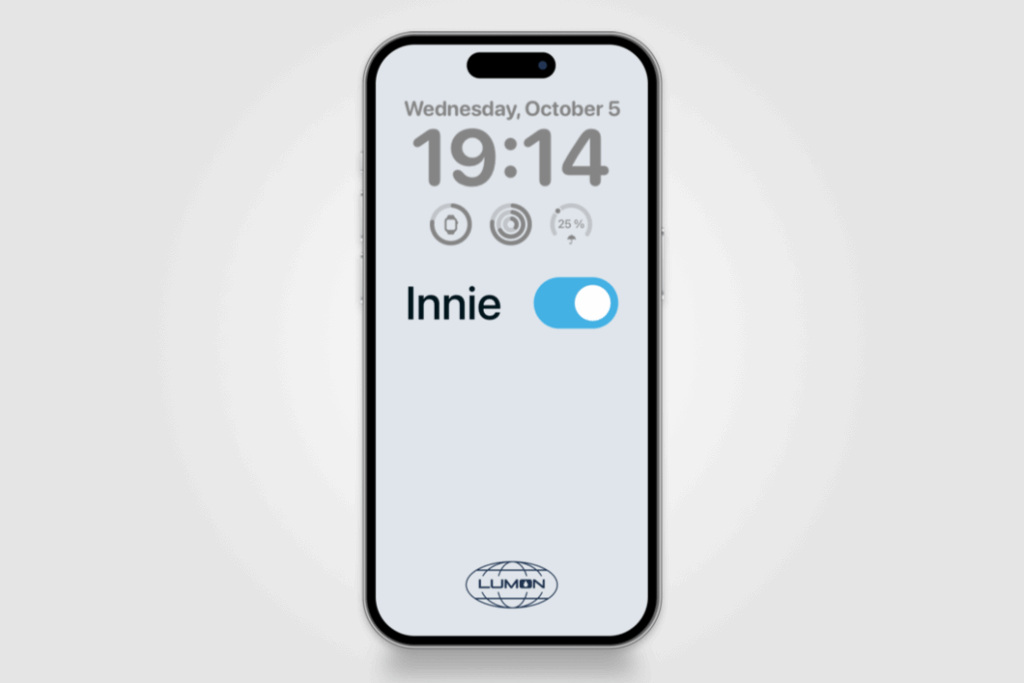 A smartphone screen shows a minimalist lock screen with a gray background. At the top, the date reads “Wednesday, October 5,” and the large time display shows “19:14.” Below the time are three circular icons representing battery, activity rings, and weather (25% chance of rain). Centered on the screen is a blue toggle switch labeled “Innie,” which is turned on. At the bottom is the Lumon Industries logo, featuring a globe outline with the word “LUMON” in the center — a reference to the TV series Severance.
